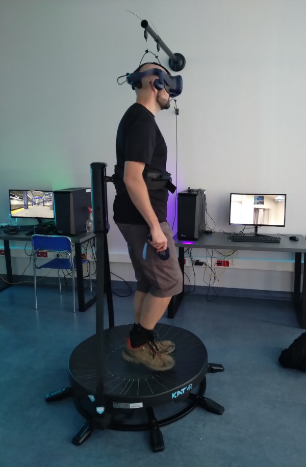 mężczyzna w laboratorium korzystający ze stanowiska VR prezentującego grę o starości. Zapięty jest w uprzęż do grania w VR, na głowie ma headset HTC vive, w rękach kontrolery, do butów przypięte są czujniki umożliwiające chodzenie w VR. 