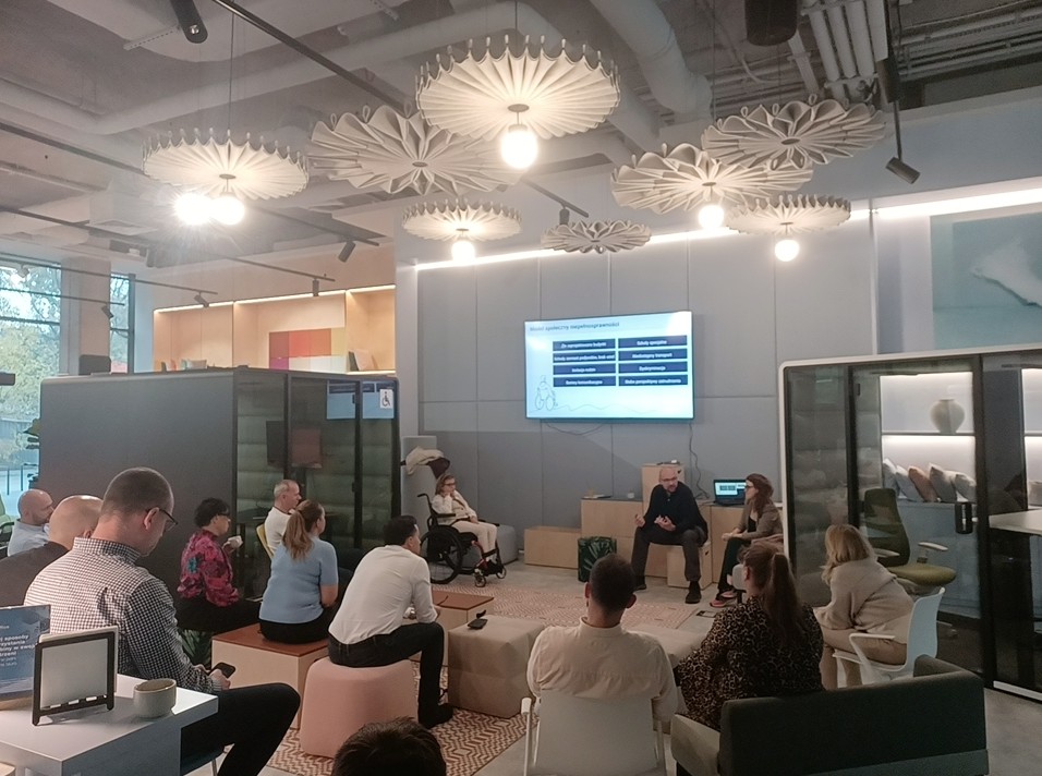 Wnętrze salonu firmy hushoffice, na lewo i na prawo kabiny wygłuszające ich produkcji. Trójka trenerów Piotr, Kasia i Magda pod obrazem z rzutnika, sala pełna ludzi.