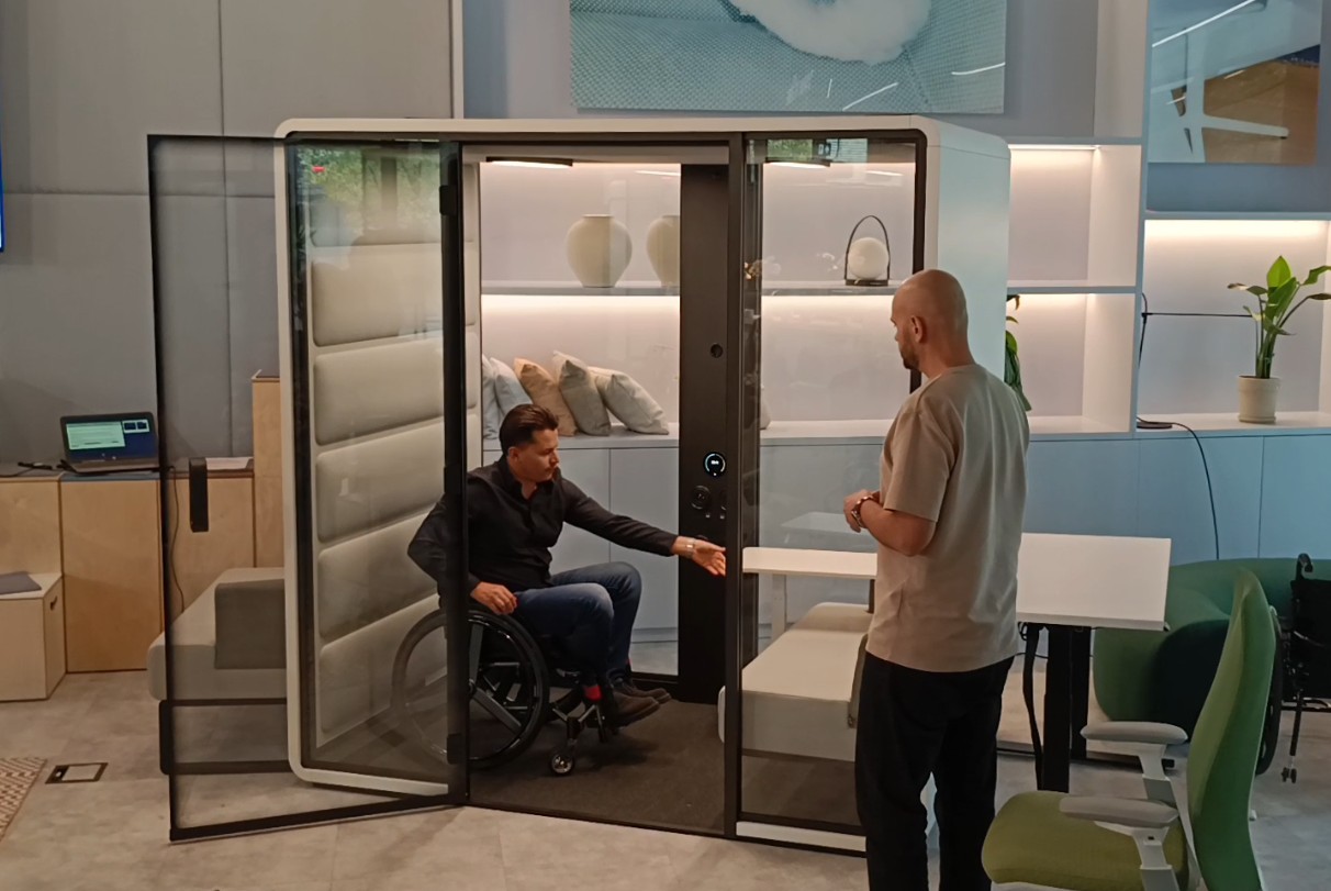 Osoba na wózku testuje ustawienie mebli wewnątrz mniejszej kabiny wygłuszającej z możliwym wjazdem dla wózków.