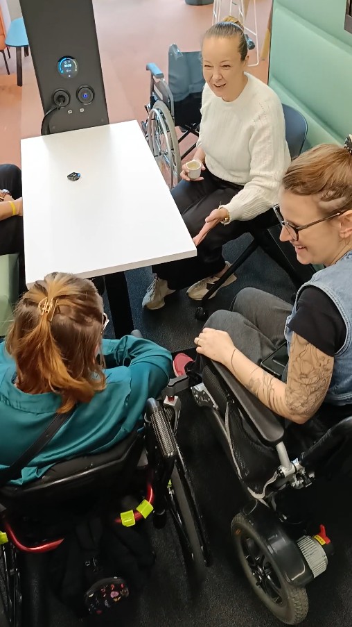 Kasia i Magda na wózkach siedzące przy stole wewnątrz kabiny wygłuszającej.