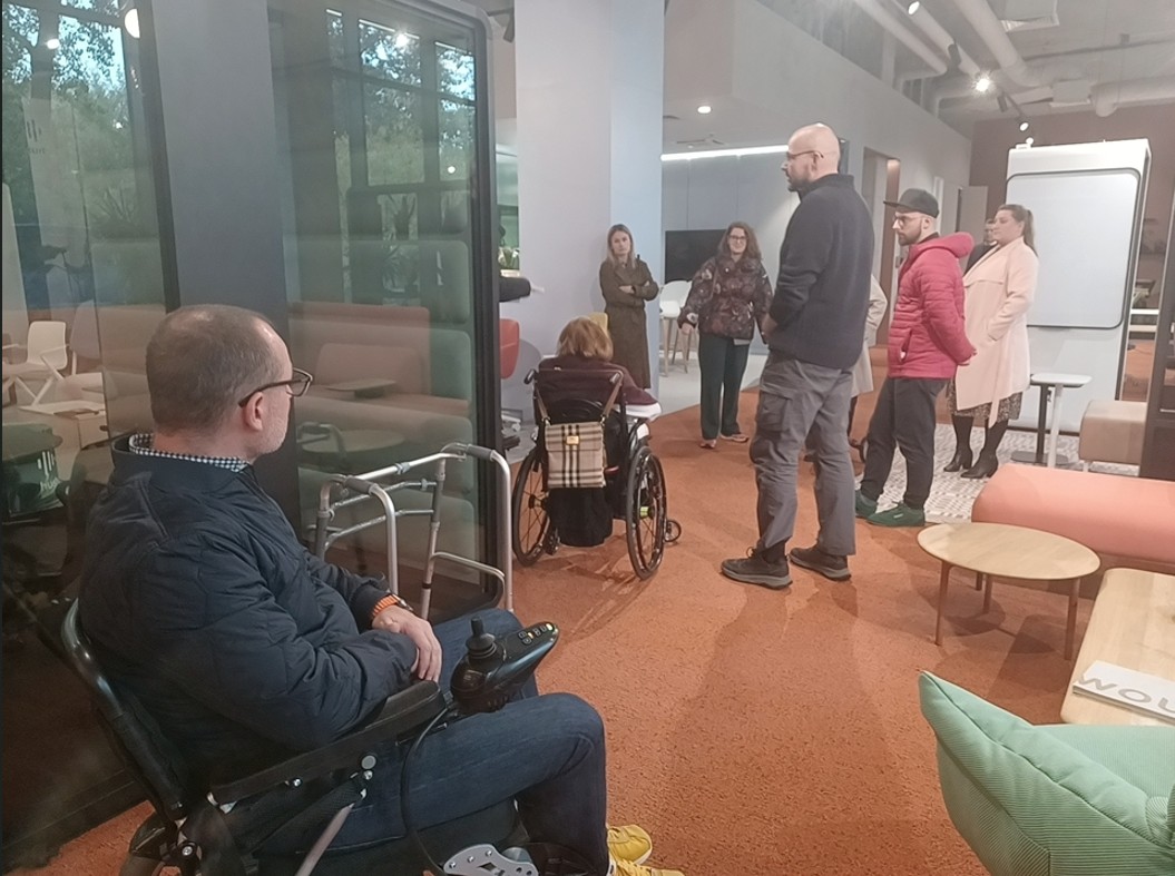 Dwie osoby na wózkach i grupa osób przygotowanych do próbnej ewakuacji budynku, w kurtkach przed wyjściem.