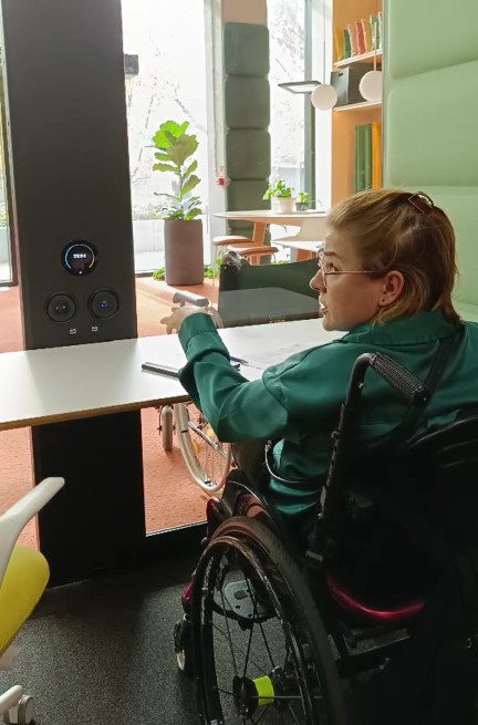 Kasia na wózku, wewnątrz kabiny, przy stole, pokazuje na zbyt daleko umieszczony dotykowy kontroler do światła i wentylacji.