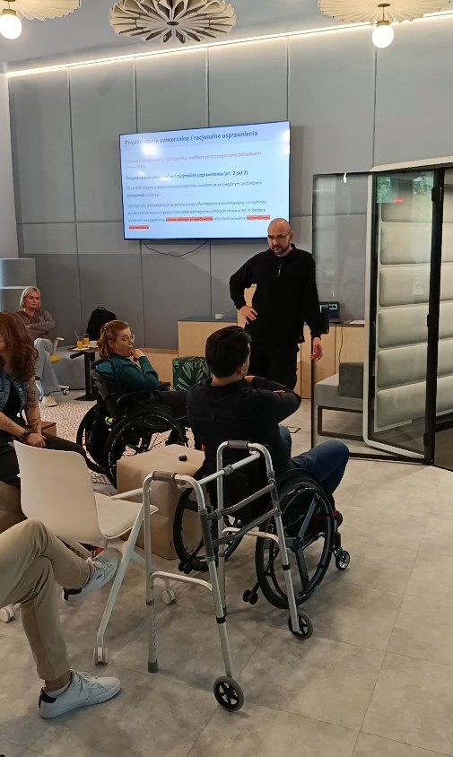 Osoby na wózkach siedzą przed wjazdem do kabiny i dyskutują, na pierwszym planie balkonik.