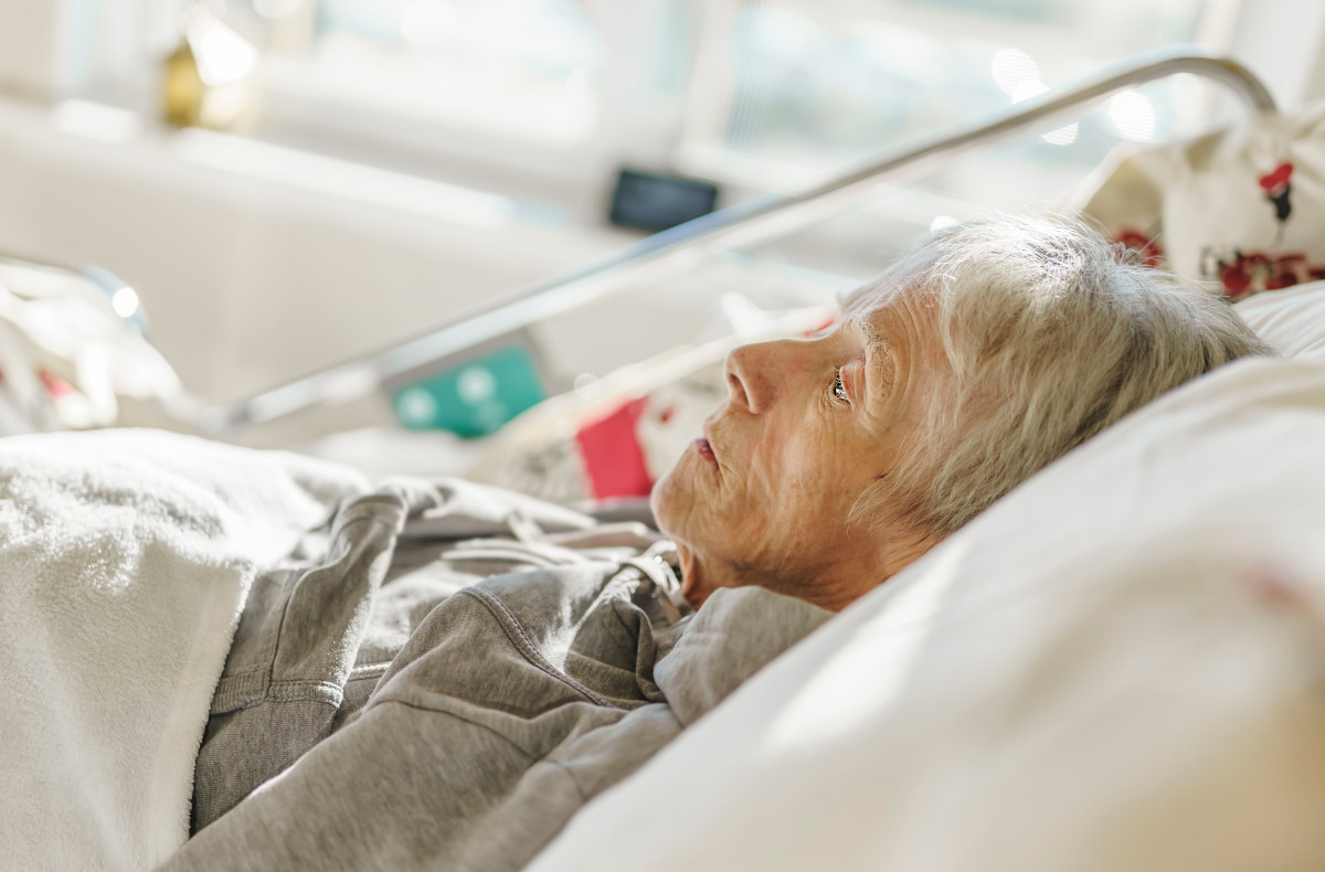 Starsza kobieta leżąca w szpilanym łóżku, patrzy przed siebie, widoczna z profilu pomarszczona twarz, usta ma zamknięte oczy otwarte
